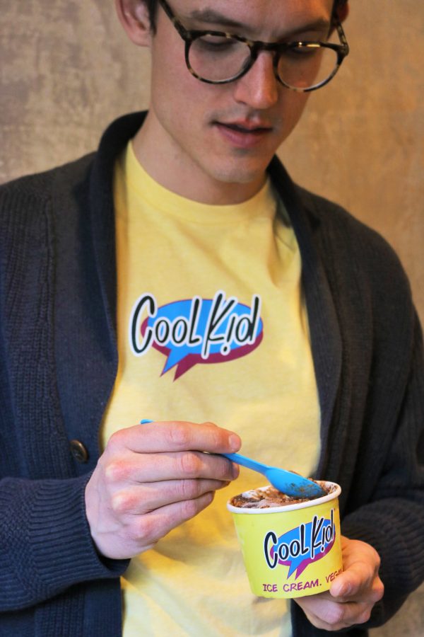 Model eating chocolate ice cream and wearing dark grey sweater over yellow Cool Kid t-shirt